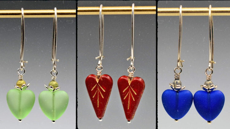 A row of three pairs of heart earrings on long silver wires. The left pair are pale green, the center are red, and the right are dark blue.
