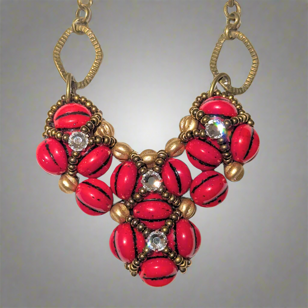 A red beaded heart pendant on a gray background. The heart is made from red beads with black lines, with an overlay of gold seed bead X-shapes with clear rhinestones in their centers.