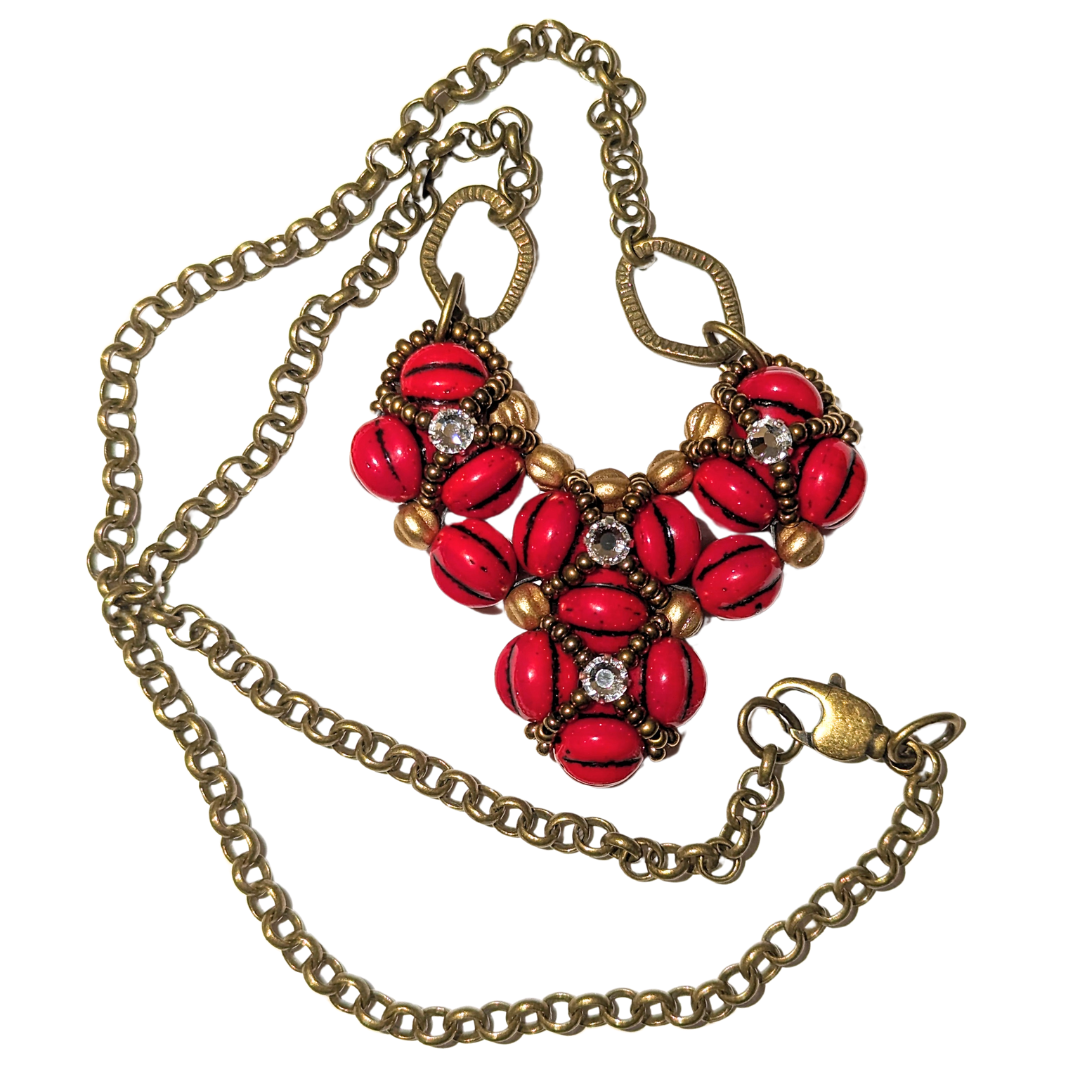 A red beaded heart pendant with a brass chain circling around iton a white background. The heart is made from red beads with black lines, with an overlay of gold seed bead X-shapes with clear rhinestones in their centers.
