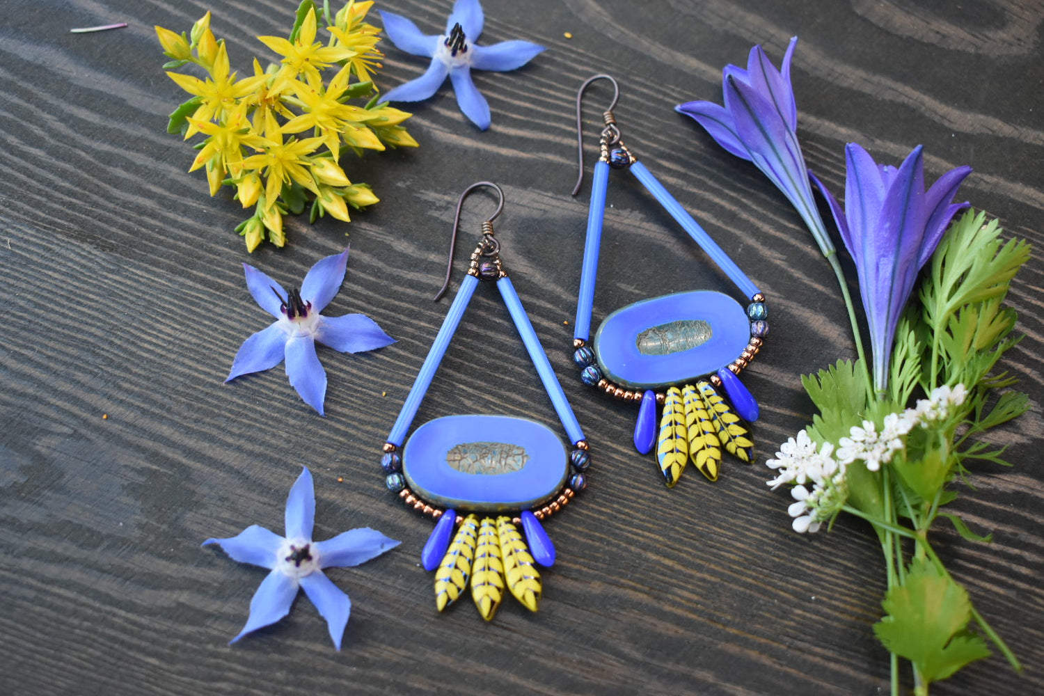 A pair of teardrop shaped earrings with large wide blue ovals in the center and a fringe of blue and yellow dagger beads lays on a wood background. There are blue and yellow flowers scattered around the earrings. 