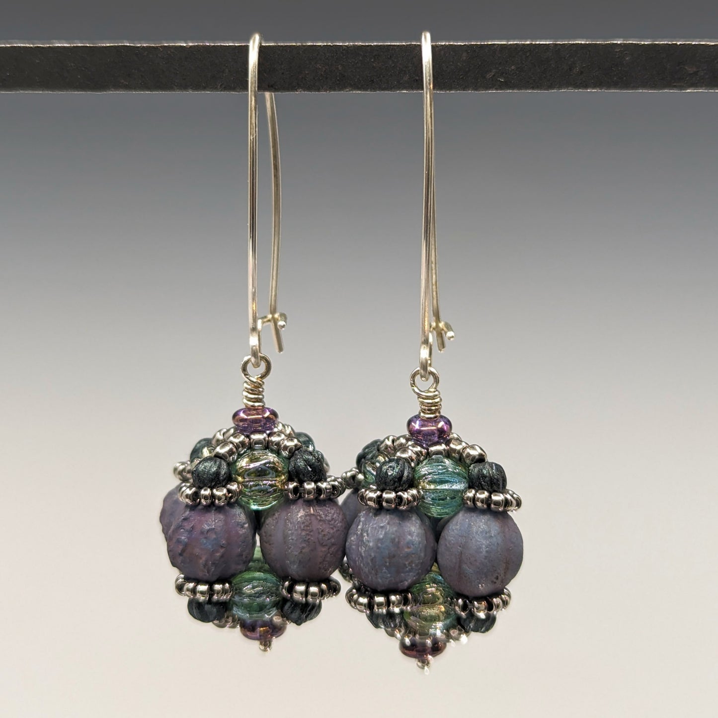 A pair of earrings hang from a black bar, against a gray background. The earrings have long silver oval wires that latch and there is a beaded ball dangling from the bottom of each earring. The beaded beads are s made from rough textured lilac beads surrounded by layers of silver and sea green beads.