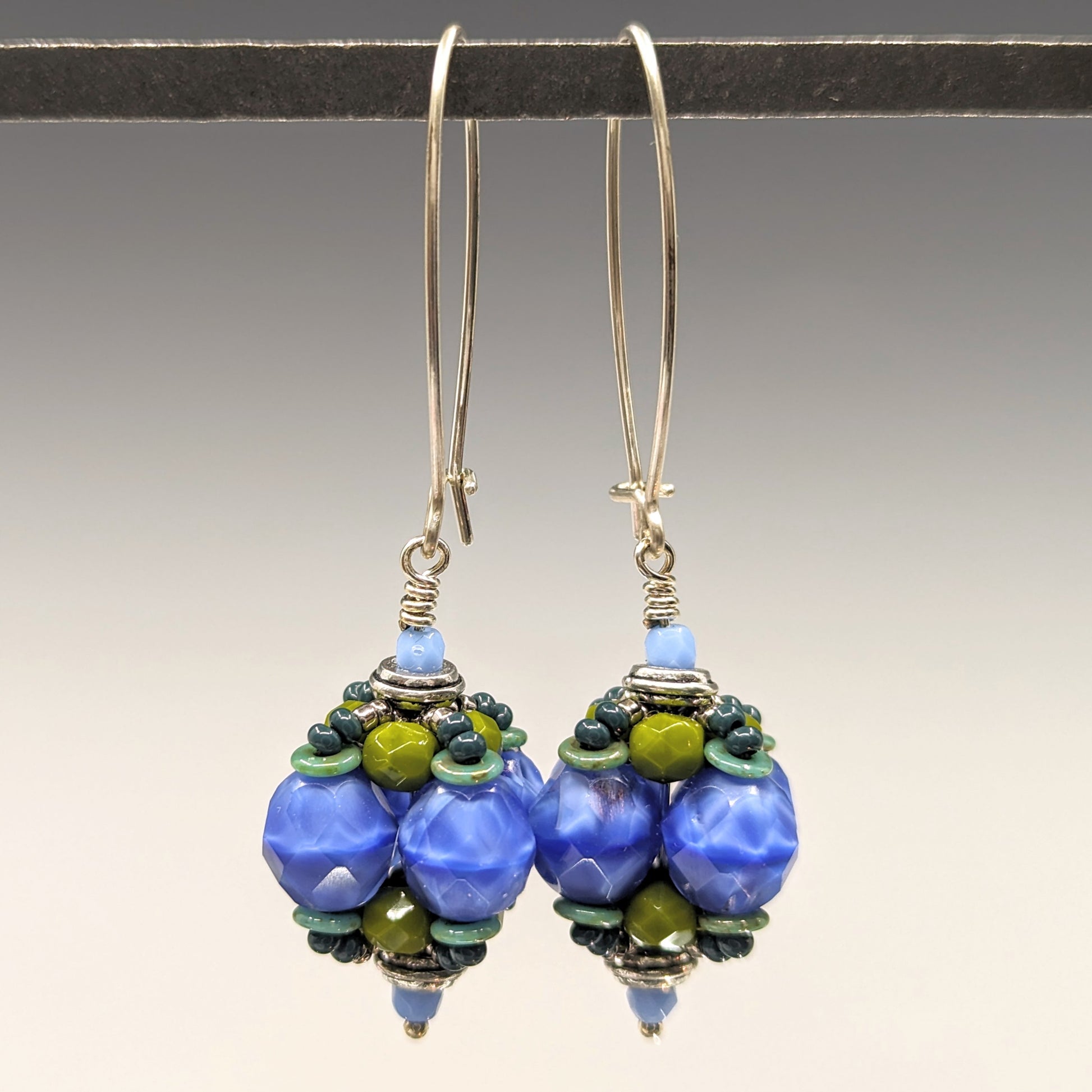A pair of earrings hang from a black bar, against a gray background. The earrings have long silver oval wires that latch and there is a beaded ball dangling from the bottom that is made from swirly blue, avocado green and speckled light turquoise beads.