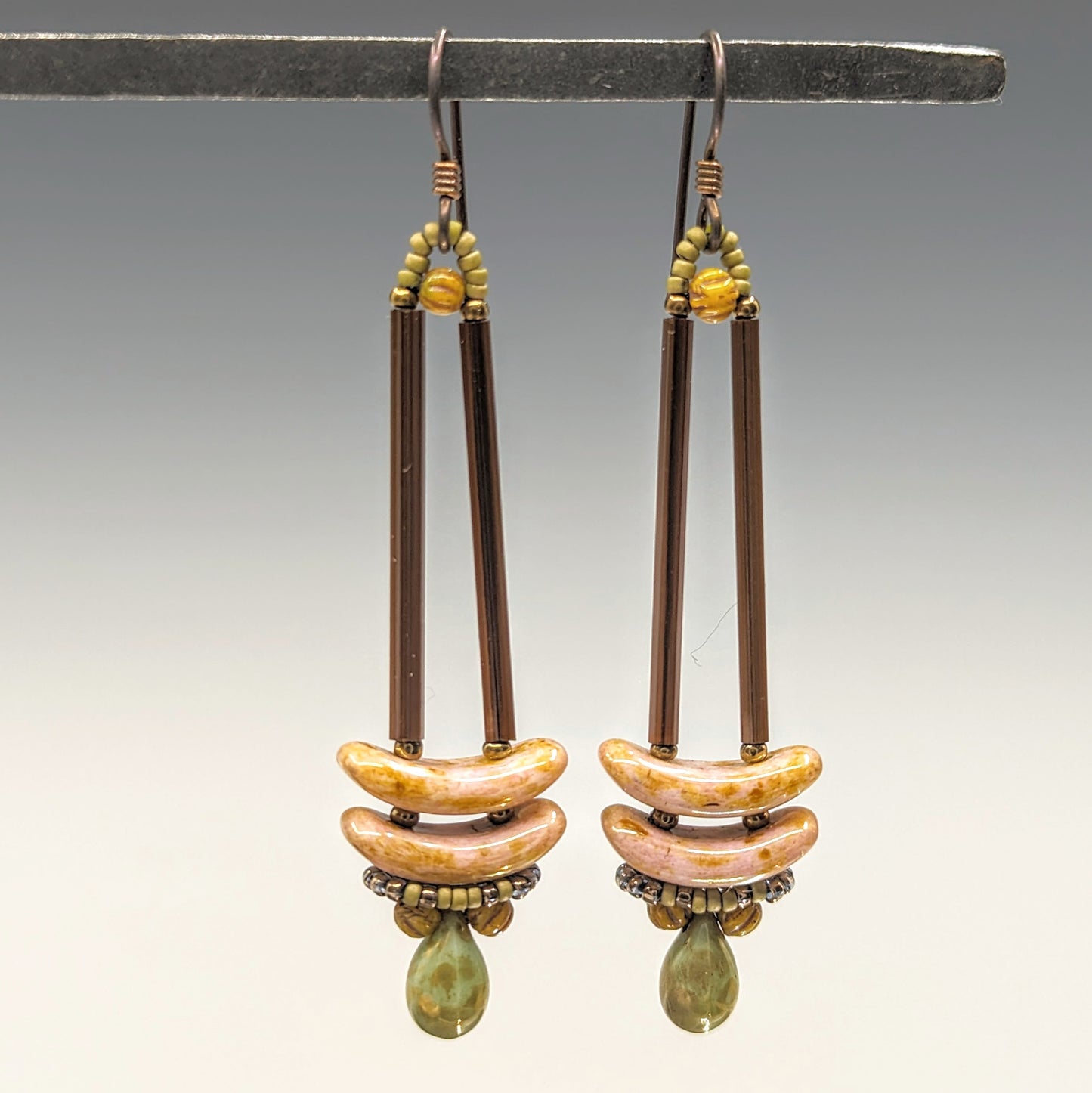 Earrings that look similar to a medicine dropper hang from a black bar. There are two long, brown beads forming narrow vertical columns ending at the top of two speckled pink stacked arc shaped beads. At the bottom is a matte speckled turquoise green drop shaped bead, surrounded by a belt of matte gold seed beads.