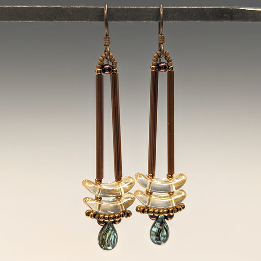 Earrings that look similar to a medicine dropper hang from  a black bar. There are two long, brown beads forming narrow vertical columns ending at the top of two clear pale yellow stacked arc shaped beads. At the bottom is a drop shaped bead made from aqua colored glass with black stripes inside of it, surrounded by a belt of gold seed beads.