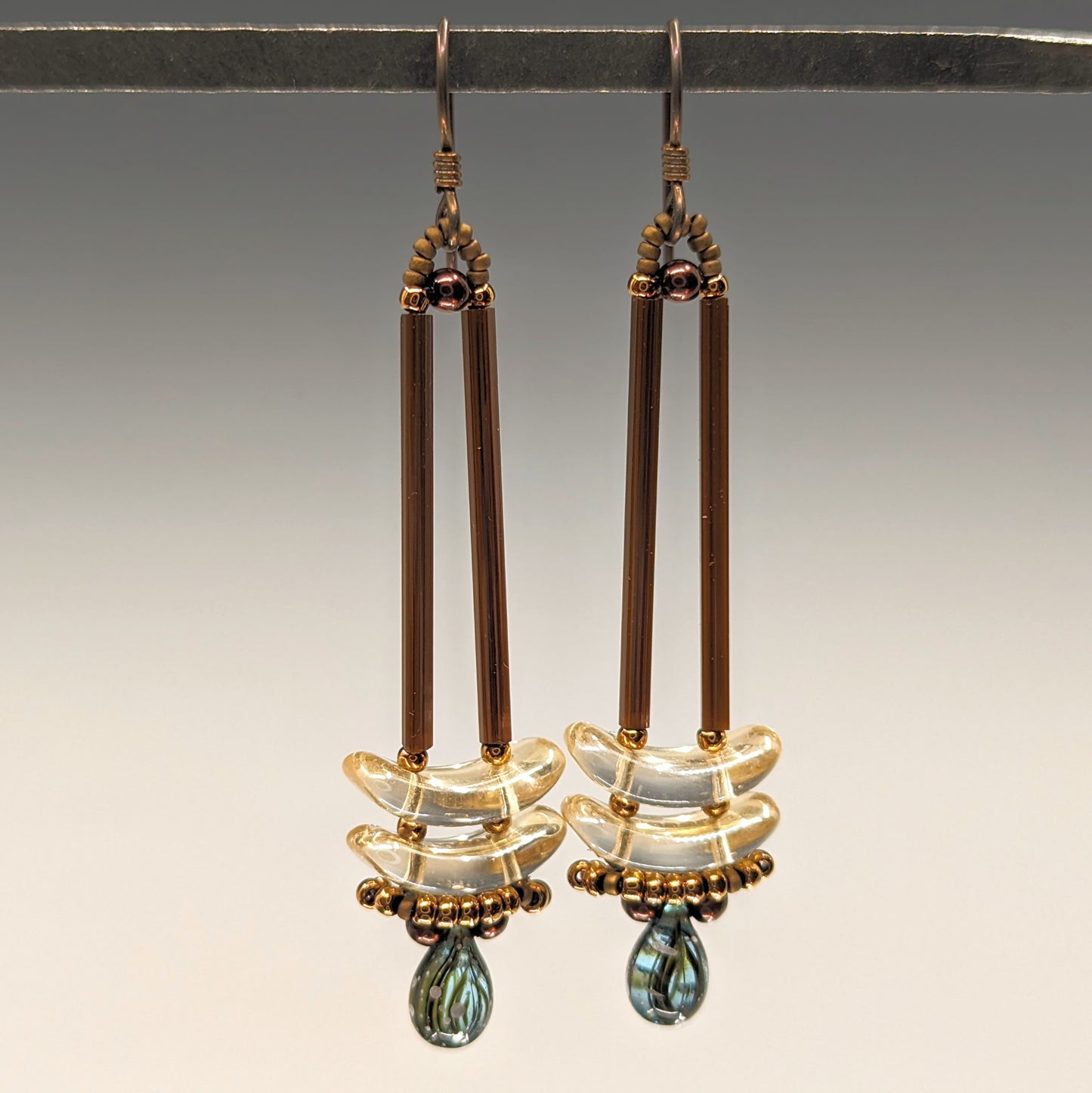Earrings that look similar to a medicine dropper hang from a black bar. There are two long, brown beads forming narrow vertical columns ending at the top of two clear pale yellow stacked arc shaped beads. At the bottom is a drop shaped bead made from aqua colored glass with black stripes inside of it, surrounded by a belt of gold seed beads.