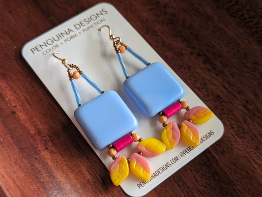 A pair of earrings on a white card lay on a reddish wood background. The earrings are have a large light blueish square suspended from gold wires. Below the square is a horizontal bright pink tube with apricot beads on either side. At the bottom is a swag of three leaf shaped beads made from variegated yellow and pastel pink glass.