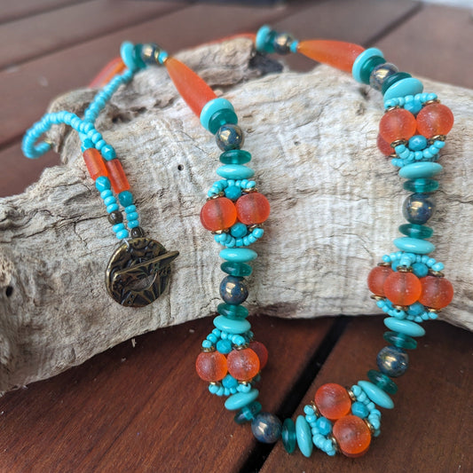 A statement necklace lays across a curved piece of driftwood. The necklace has frosted orange  beads alternating with dark to light shades of turquoise.  At the bottom of the necklace, there are five clusters of beads that have been woven into an orb. 