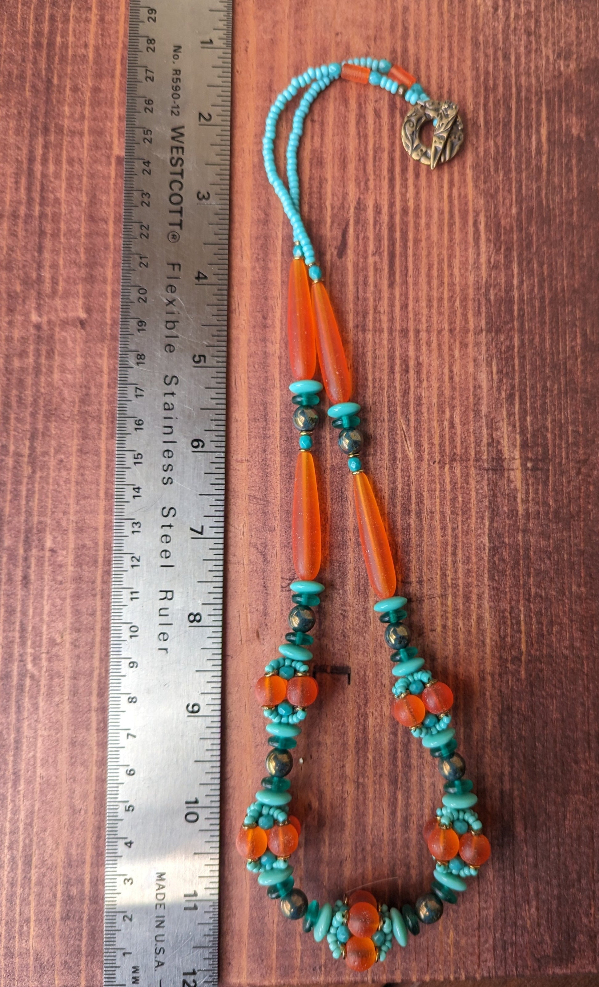 A statement necklace, photographed from above, on a reddish wood board. A twelve inch ruler lays next to the necklace. The necklace has frosted orange beads alternating with dark to light shades of turquoise. At the bottom of the necklace, there are five clusters of beads that have been woven into an orb.