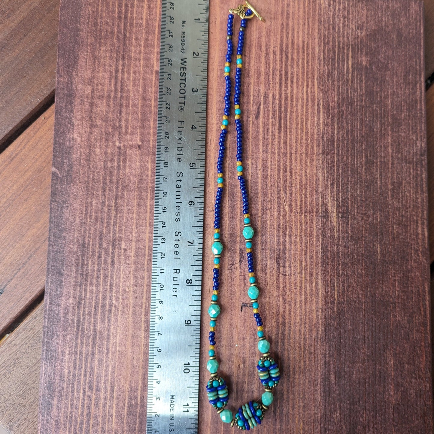A royal blue and turquoise necklace laying next to a metal ruler on a reddish wood board. The necklace has three clusters of beaded spheres. 