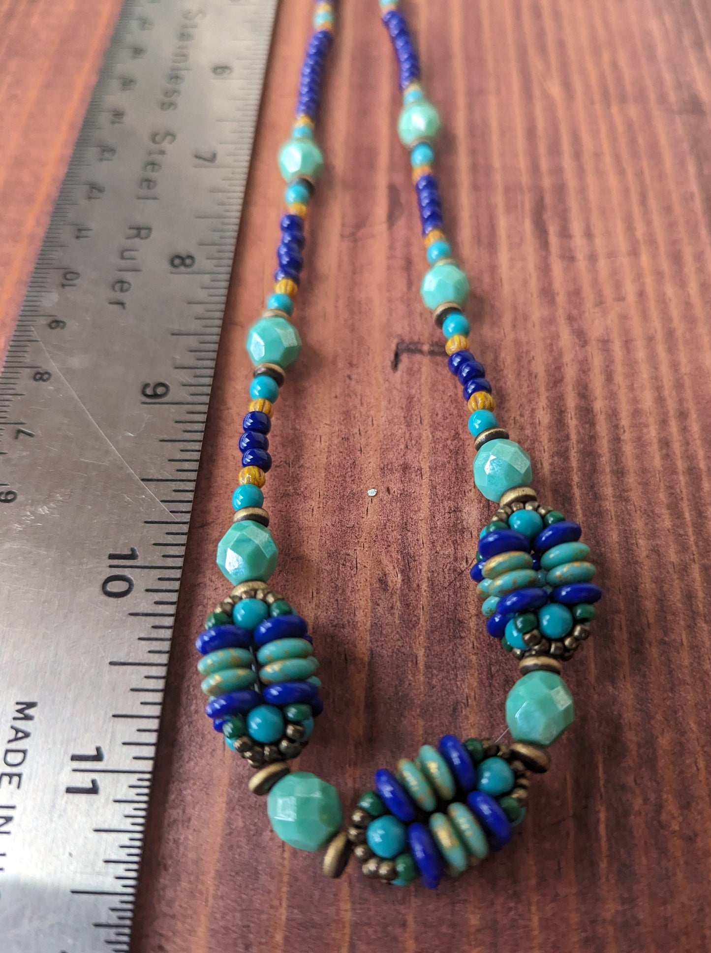 A royal blue and turquoise necklace laying next to a metal ruler on a reddish wood board. The necklace has three clusters of beaded spheres.  The  image is photographed as an angle that makes the bottom of the necklace and ruler appear larger. 