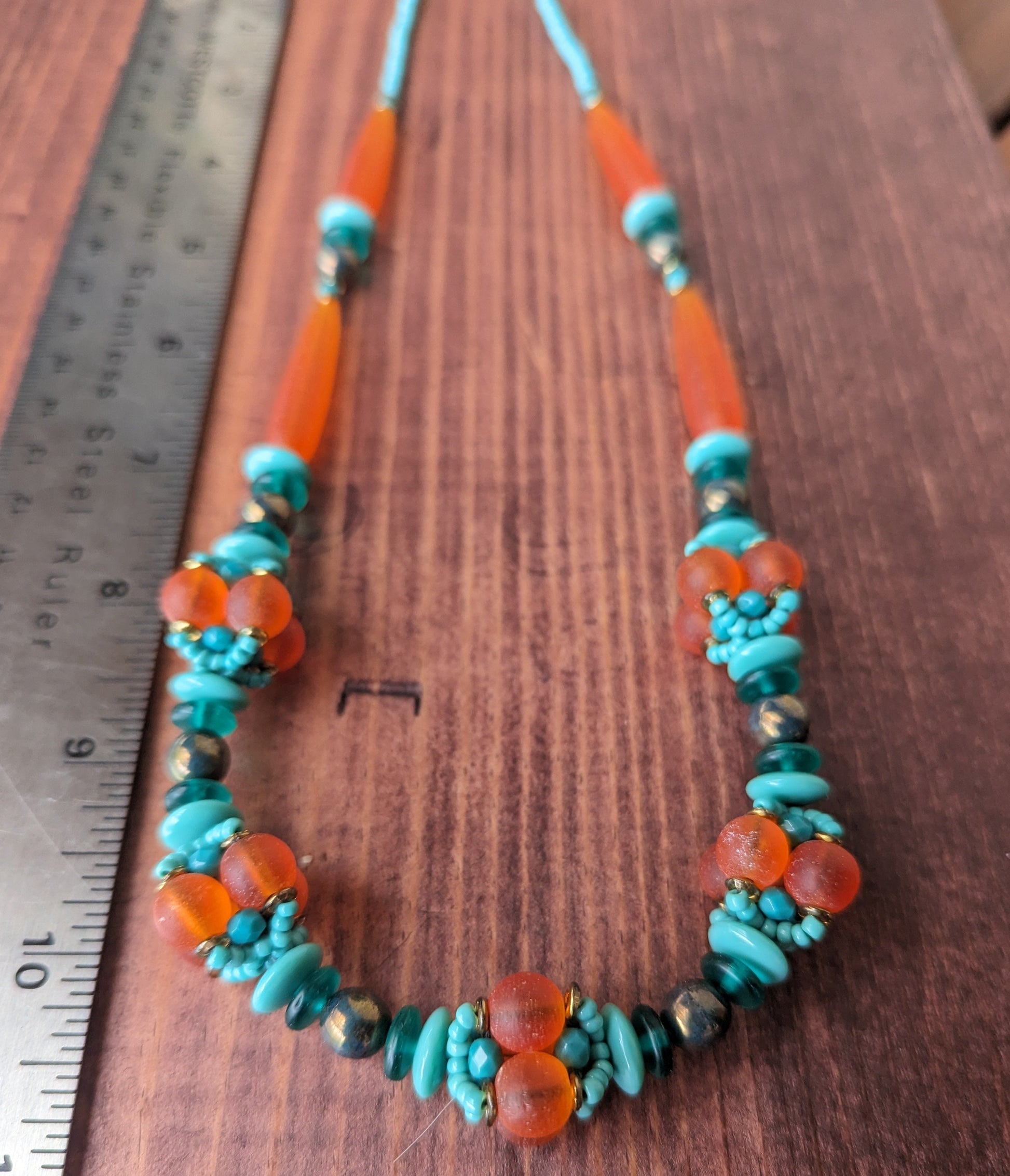 A statement necklace lays on a reddish wood board, next to a metal ruler. The necklace has frosted orange  beads alternating with dark to light shades of turquoise.  At the bottom of the necklace, there are five clusters of beads that have been woven into an orb. 