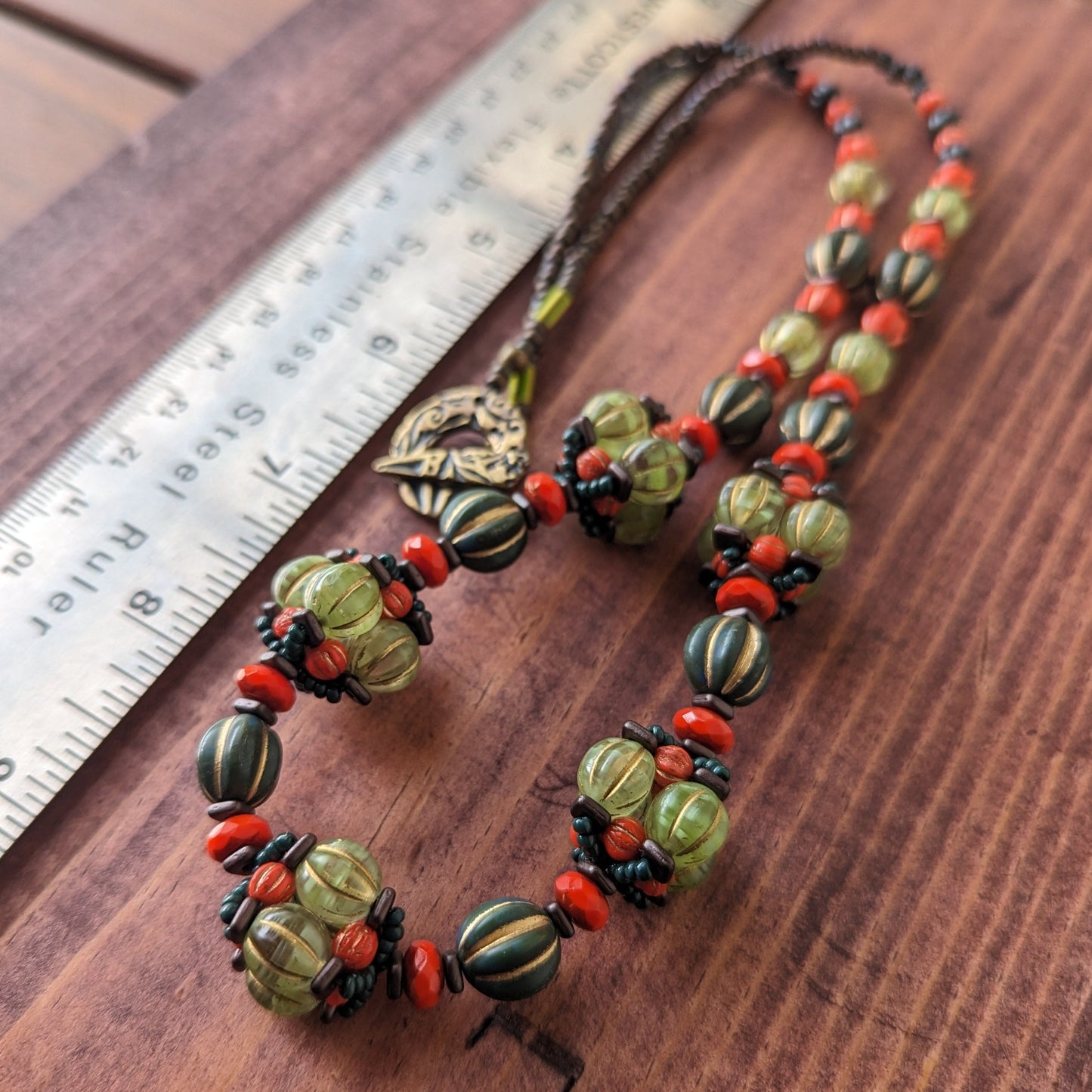 A beaded necklace made with alternating dark green and coral beads, with five pale green and orange beaded beads added into the pattern at the front. The necklace is laying on a reddish wooden board next to a ruler.