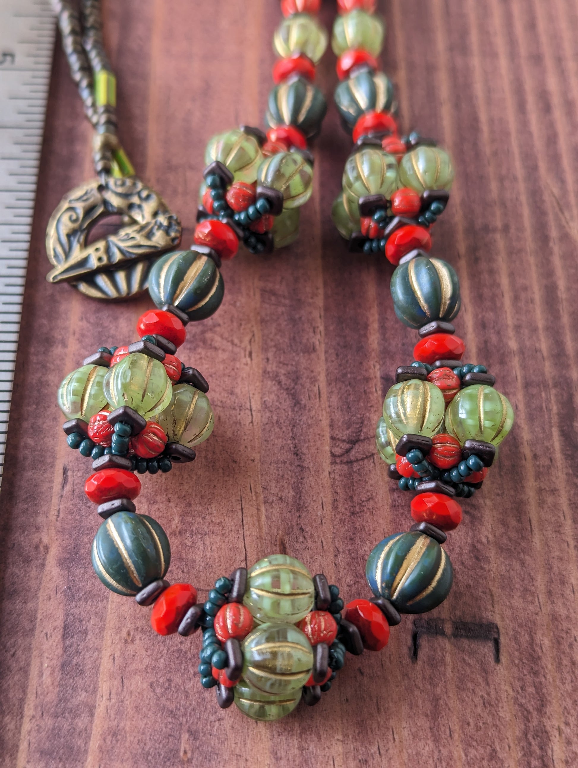 A close up of a necklace with five pale green and orange beaded beads alternating with dark green and coral beads. The necklace is laying on a reddish wooden board, with just the hash-marked edge of a metal ruler visible on the left. 