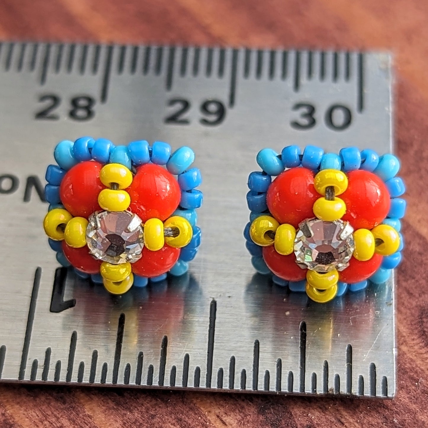 Square stud earrings formed from four red beads with a clear rhinestone held in place by an x-shape of yellow seed beads. The outside of the earrings is outlined in medium blue seed beads. The earrings are resting on a silver metal ruler showing they are about 3/8ths inch wide.