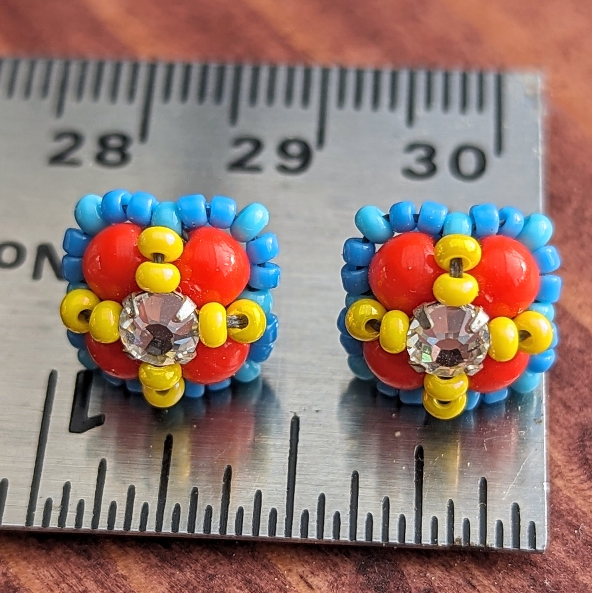 Square stud earrings formed from four red beads with a clear rhinestone held in place by an x-shape of yellow seed beads. The outside of the earrings is outlined in medium blue seed beads. The earrings are resting on a silver metal ruler showing they are about 3/8ths inch wide.