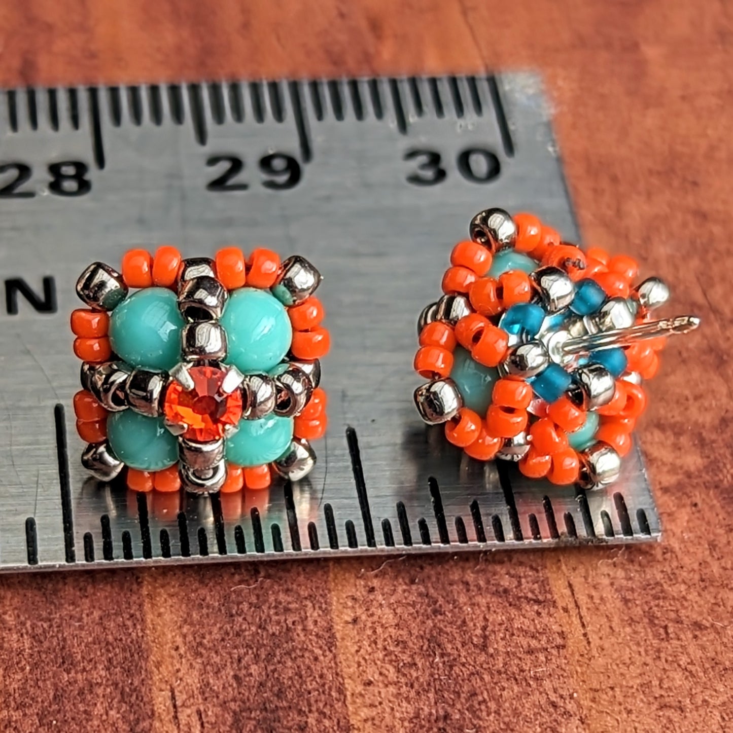 A pair of square stud earrings resting on a metal ruler to show that they are a little less than a half inch across. One stud is positioned to show the back, which has a net of small beads holding the finding in place. The forward facing earring is formed from four turquoise beads with an orange rhinestone. The rhinestone is held in place by an x-shape of silver seed beads. The outside of the earrings is outlined in orange seed beads.