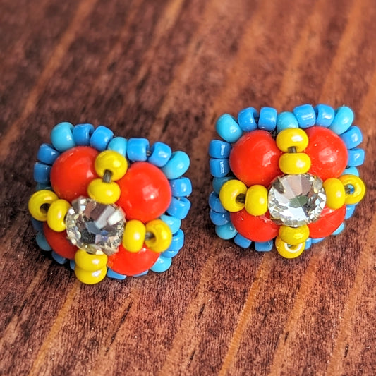 Square stud earrings formed from four red beads with a clear rhinestone held in place by an x-shape of yellow seed beads. The outside of the earrings is outlined in medium blue seed beads.