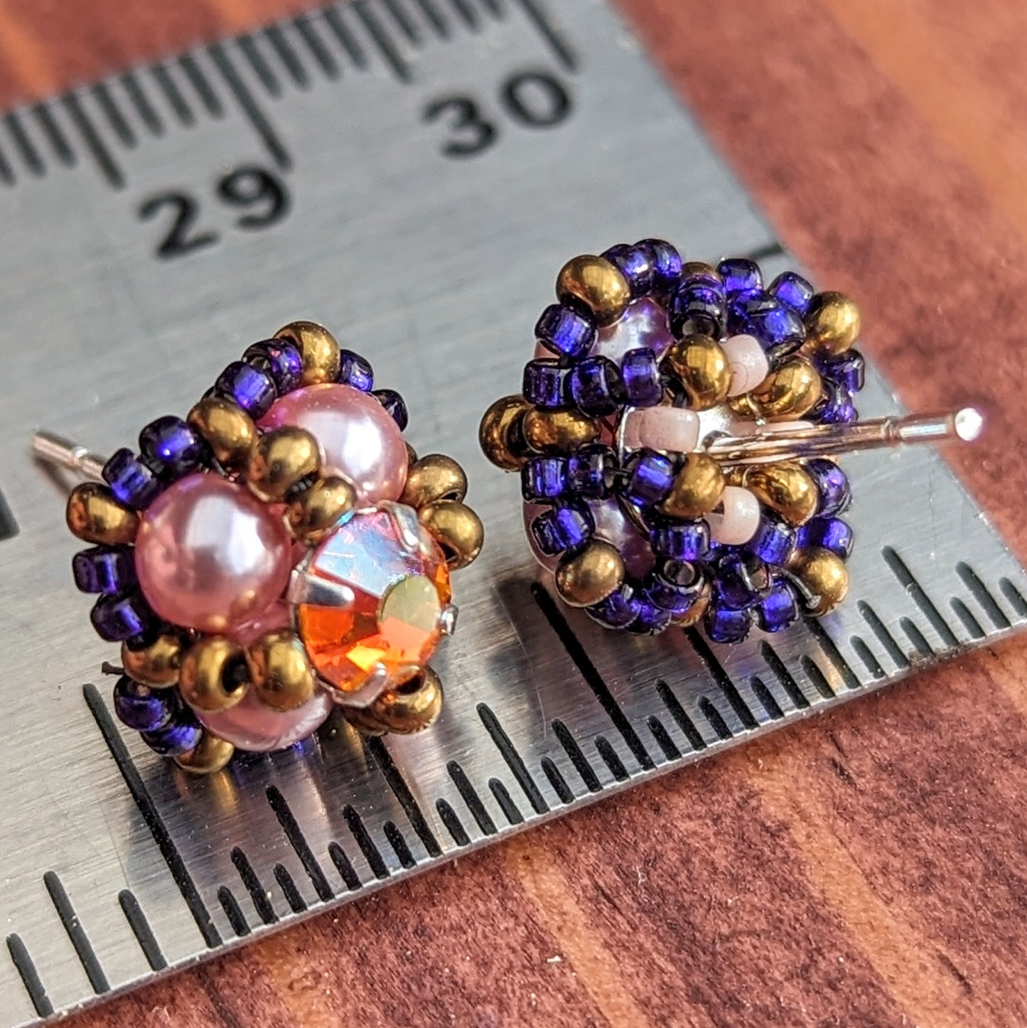 Square stud earrings resting on a metal ruler to show that they are a little less than a half inch across. The studs are placed with one cacing forwards and one showing the back, which has a net of small beads holding the finding in place. The forward facing earring is formed from pearly pale pink beads with a golden-orange and rainbow flash rhinestone. The rhinestone is held in place by an x-shape of antique gold seed beads. The outside of the earrings is outlined in dark electric purple seed beads.