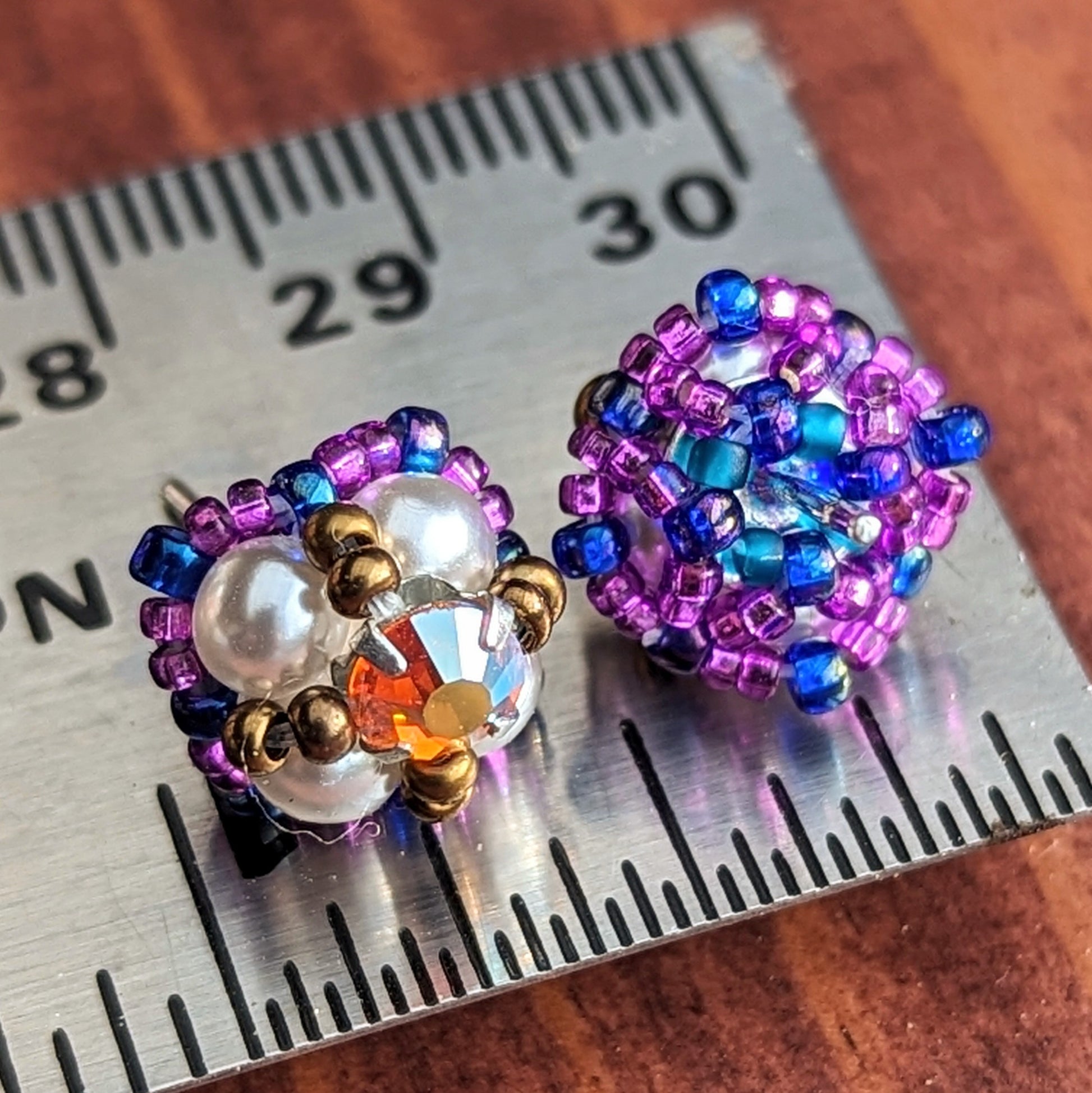 A pair of square stud earrings resting on a metal ruler to show that they are a little less than a half inch across. The studs are placed with one facing forwards and one showing the back, which has a net of small beads holding the finding in place. The forward-facing earring has four pearly white beads with a golden-orange rhinestone. The rhinestone is held in place by an x-shape of antique gold seed beads. The outside of the earrings is outlined in grape purple and dark electric purple seed beads.
