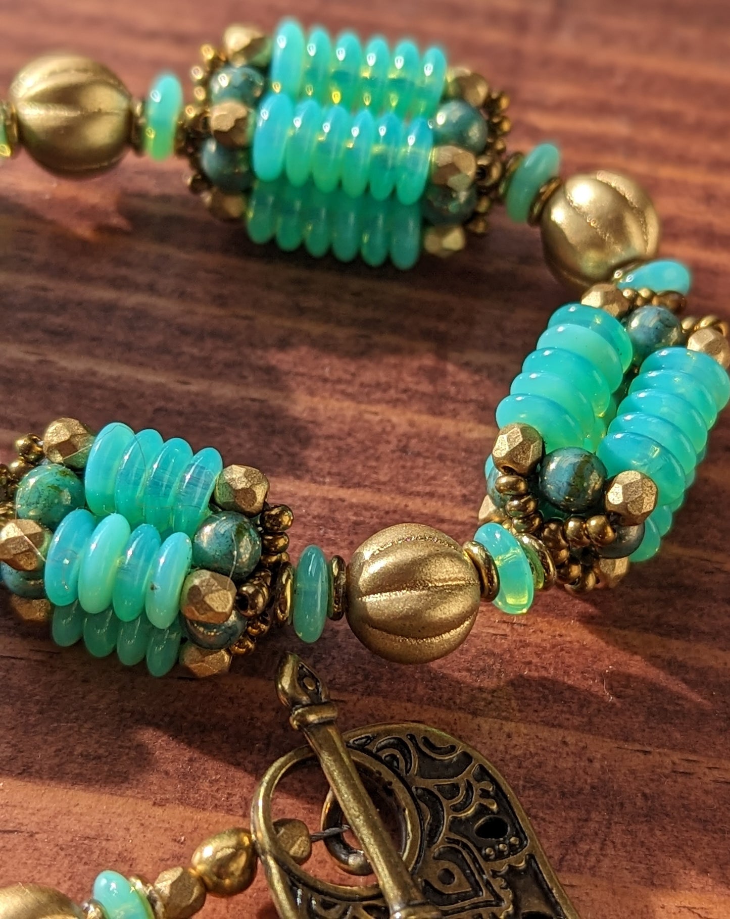 A close up of three beaded beads with columns of light opalescent seafoam Czech glass discs with lustered teal accents. Between each beaded bead is a gold melon bead. At the lower left side there is a delicately patterned teardrop shaped brass toggle clasp.