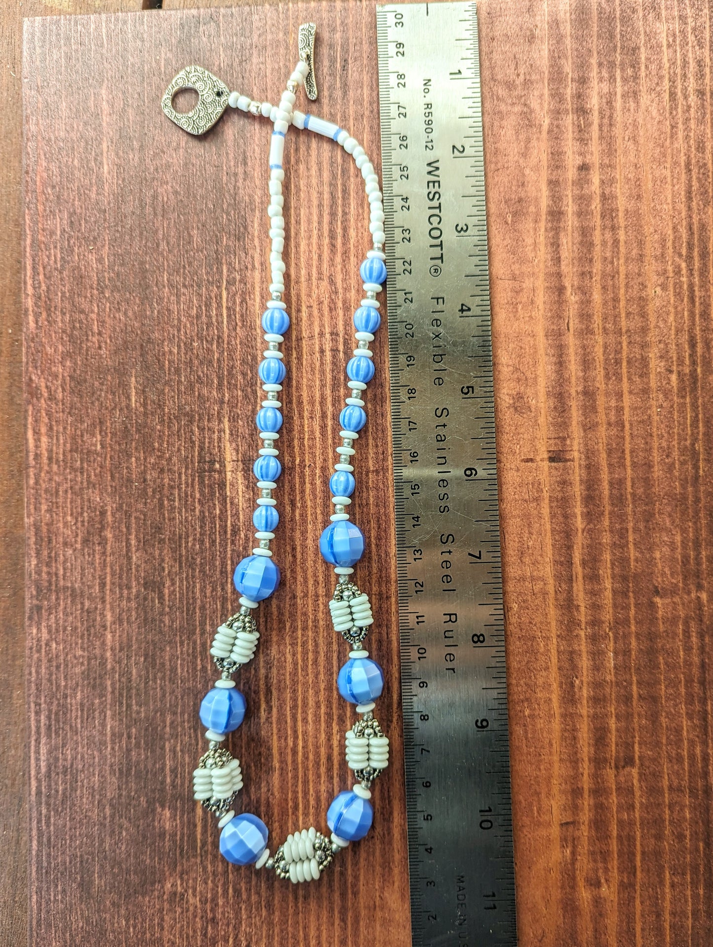 A blue, white and silver necklace is alongside a metal ruler, atop a reddish wood board. The necklace has white beads and a silver toggle clasp at the back and then graduated blue beads leading to an alternating pattern of large blue beads and beaded beads that are comprised of four columns of white discs capped by silver beads.
