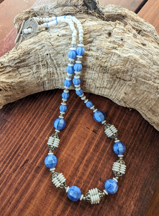 A blue, white and silver necklace is draped across a piece of driftwood. The necklace has white beads and a silver toggle clasp at the back and then graduated blue beads leading to an alternating pattern of large blue beads and beaded beads that are comprised of four columns of white discs capped by silver beads.