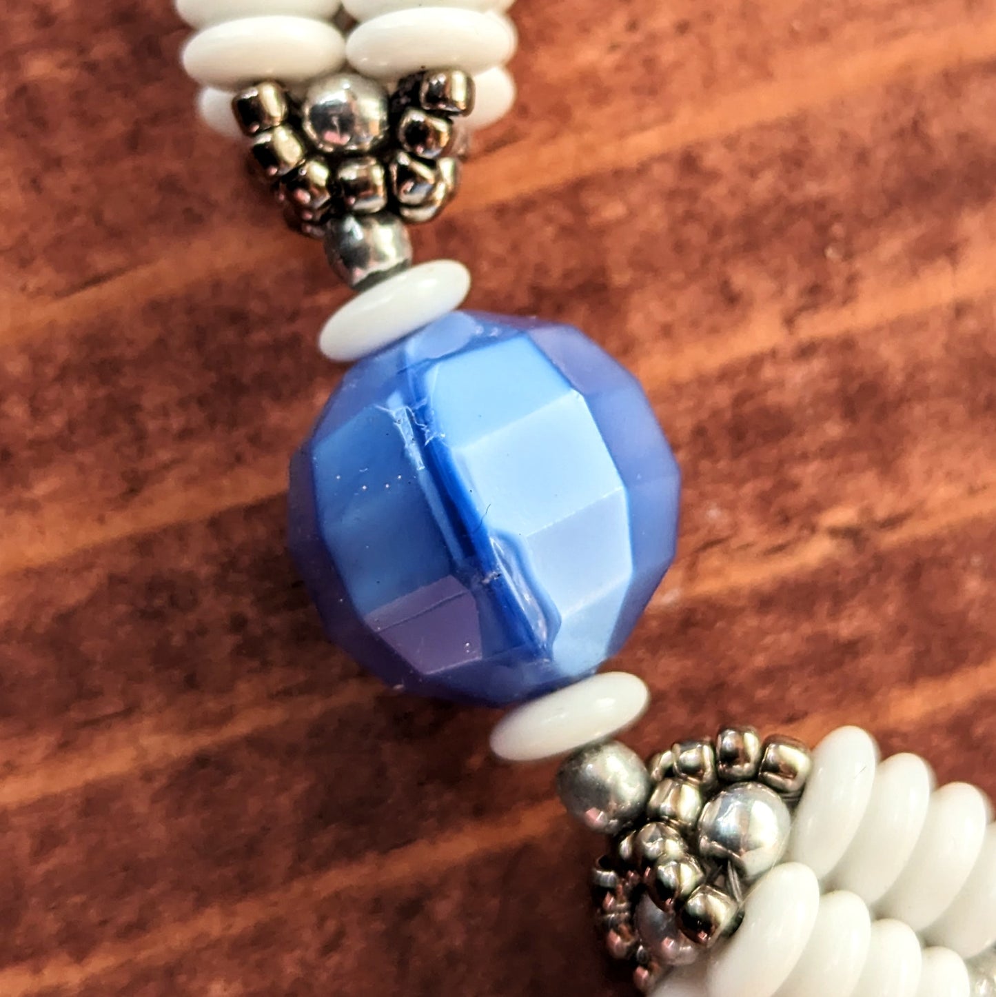 A close up of a large blue bead with white discs and a white and silver beaded bead on either side. The blue bead has soft color variations, rectangular facets, and along one vertical facet, a dark blue mold line and small  chip are visible.