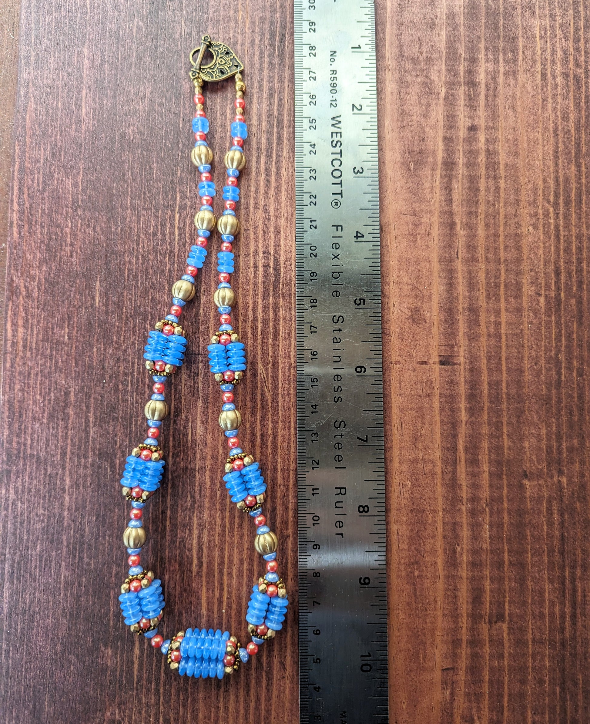 A necklace with bright opal blue, gold, and orangey red beads is laid out alongside a metal ruler on a reddish wood board. A teardrop shaped brass toggle clasp is visible. The necklace has nine beaded beads that are comprised of four columns of blue discs and capped with gold and red beads