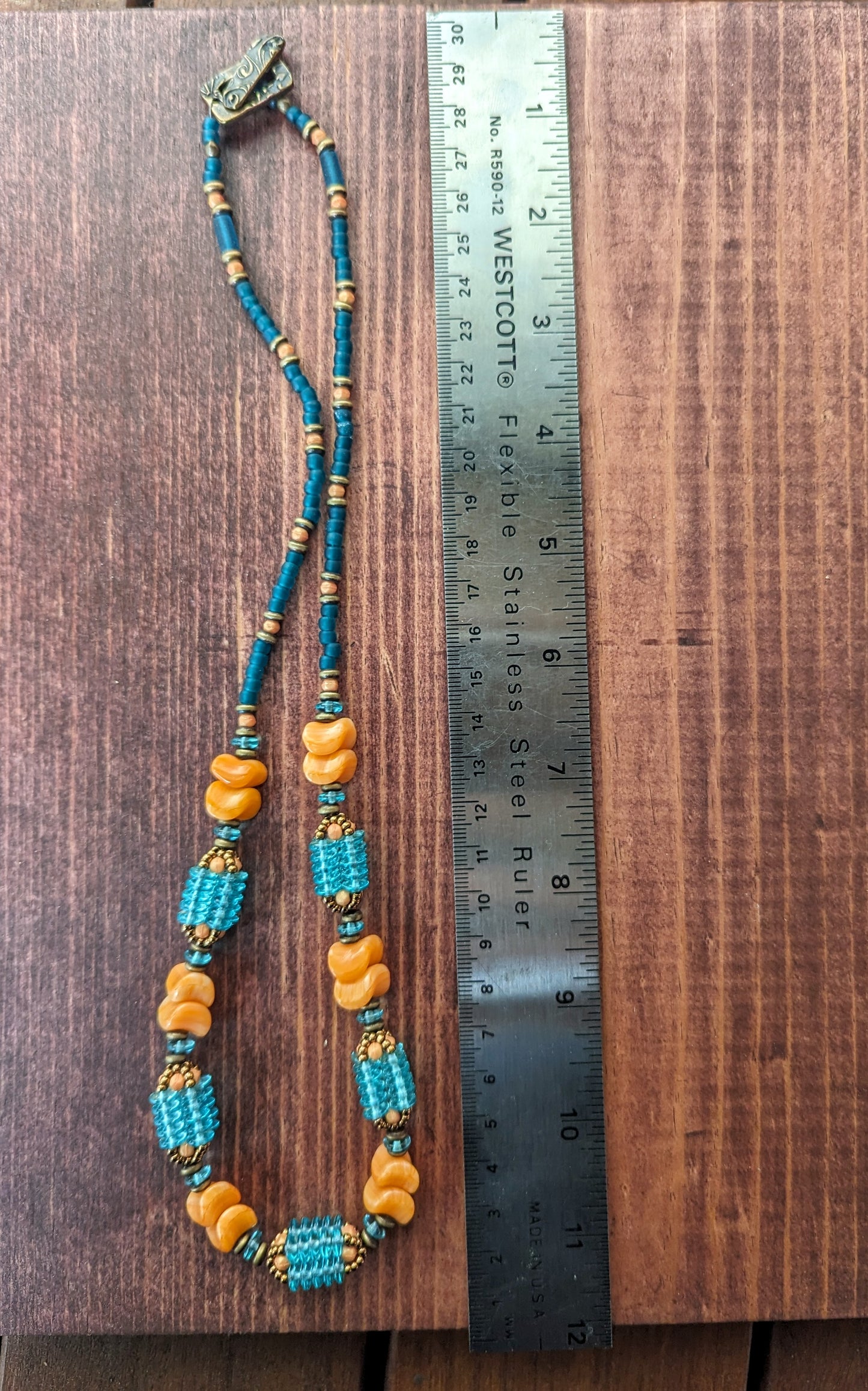 A light orange and transparent teal necklace laid out in a loop alongside a metal ruler. The necklace has orange beads shaped like waves and teal beaded beads that are made up of four columns of teal discs.