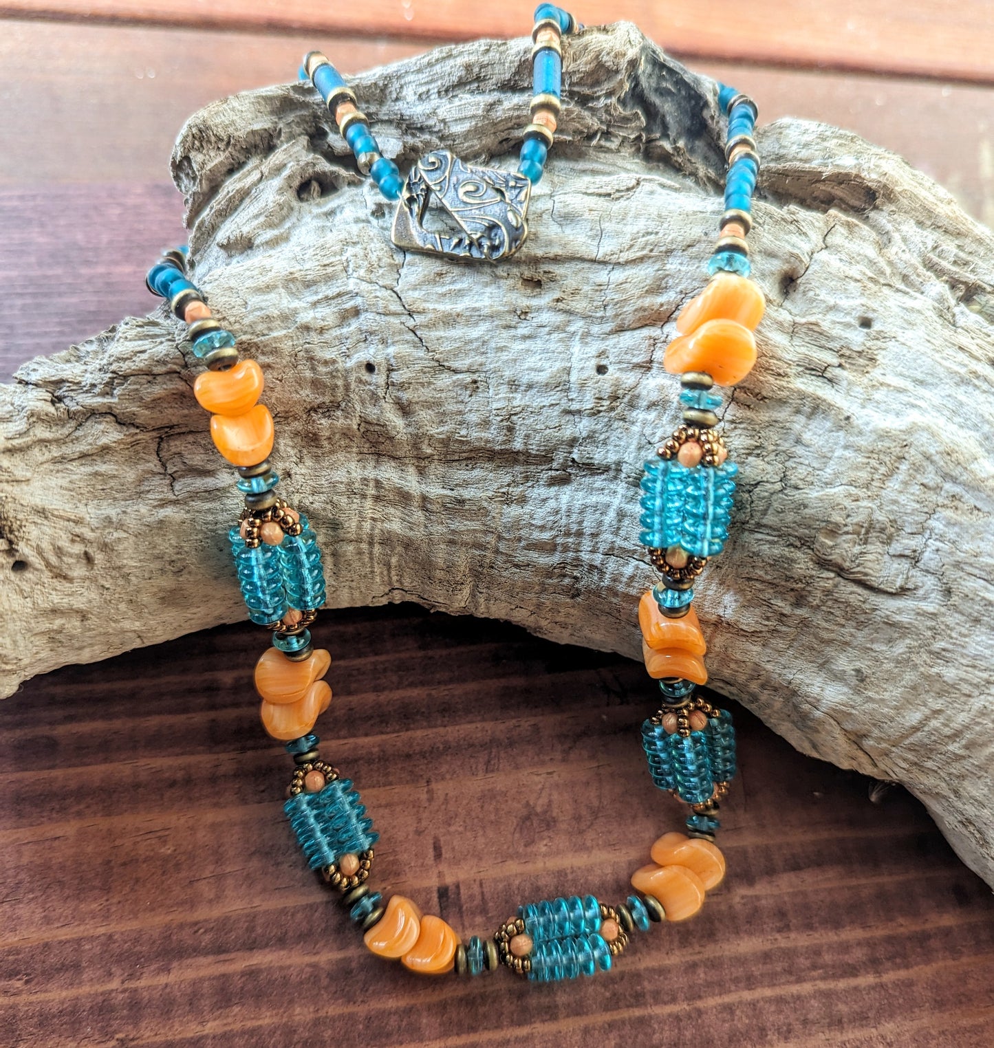 A light orange and transparent teal necklace draped across a curved gray piece of driftwood. The necklace has orange beads shaped like waves and teal beaded beads that are made up of four columns of teal discs.