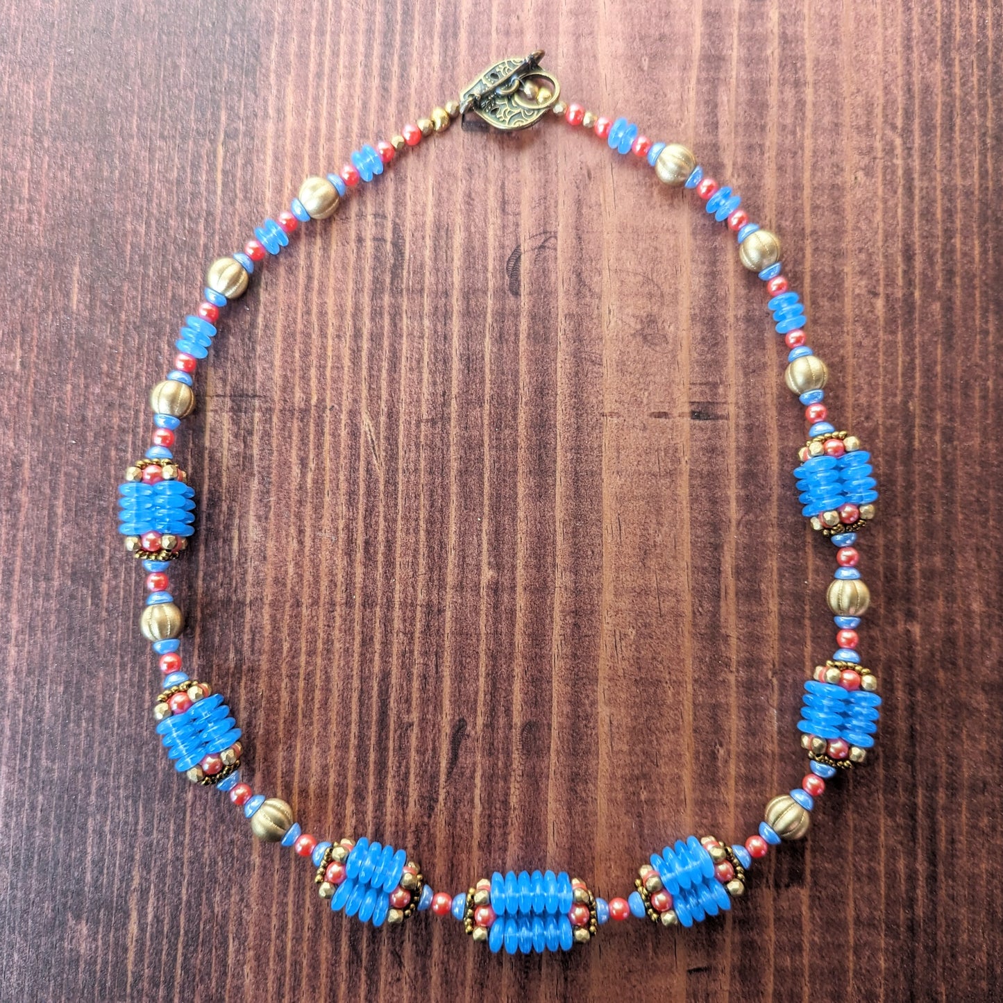 A necklace with bright opal blue, gold, and orangey red beads is laid out in a  on circlea reddish wood board. A teardrop shaped brass toggle clasp is visible. The necklace has nine beaded beads that are comprised of four columns of blue discs and capped with gold and red beads