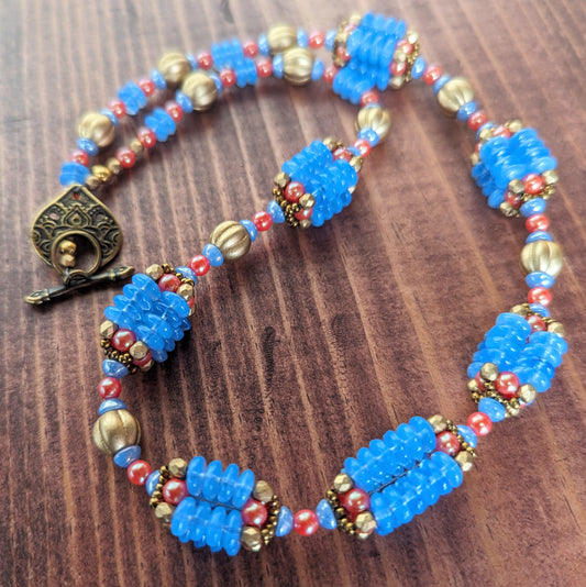 A necklace with bright opal blue, gold, and orangey red beads is draped in a loop on a reddish wood board.  A teardrop shaped brass toggle clasp is visible. The necklace has nine beaded beads that are comprised of four columns of blue discs and capped with gold and red beads.