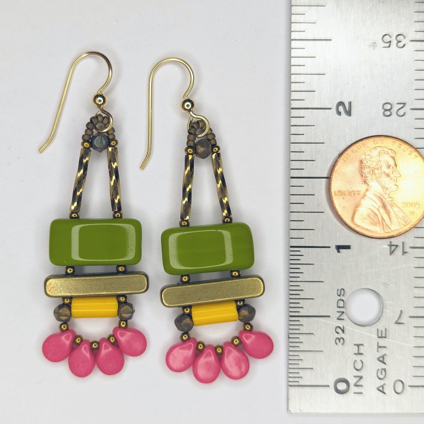 Long earrings laying next to a ruler with a penny on it. The earrings each have a wide avocado green rectangle stacked on top of a brass colored bar. Under the bar there is a light orange tube and a swag of four medium rose pink petal beads.