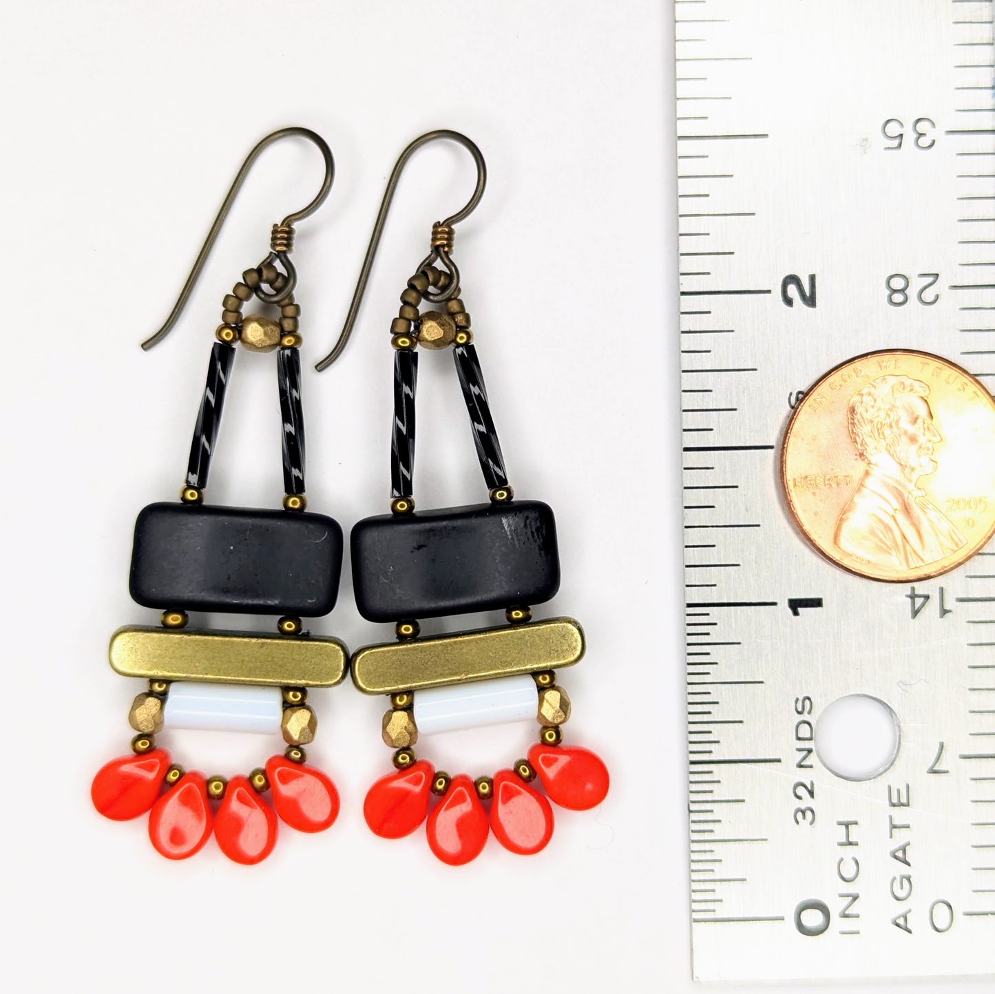 Long earrings laying next to a ruler with a penny on it. Each earring has a wide matte black rectangle stacked on top of a brass colored bar. Under the bar there is a white tube and a swag of four warm red petal beads.