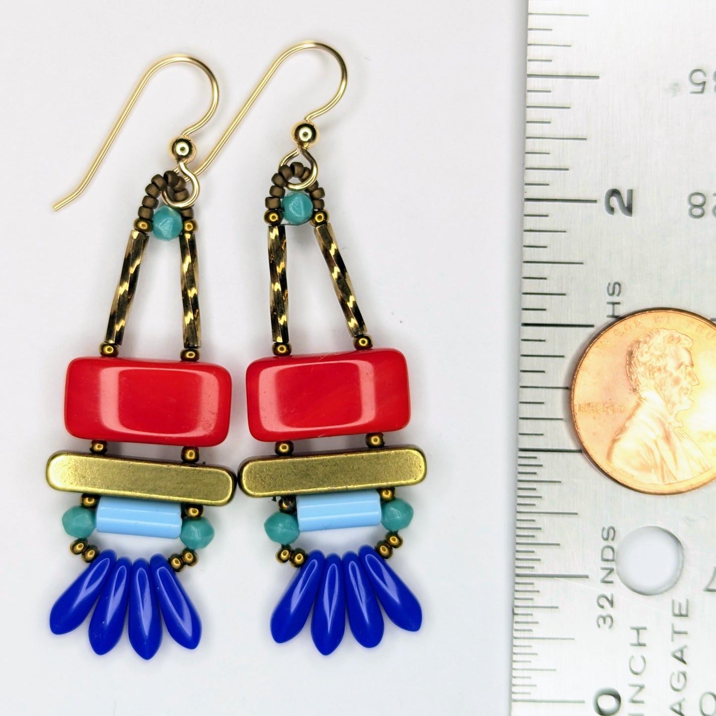 Long earrings laying next to a ruler with a penny on it. The earrings each have a wide red rectangle stacked on top of a brass colored bar. Under the bar there is a light blue tube and a swag of four small blue dagger beads.