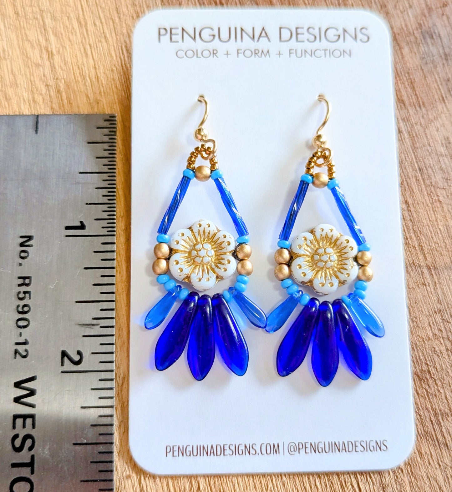 Beaded earrings with a white flower surrounded by blue and gold beads on a Penguina Designs card with a ruler for scale.