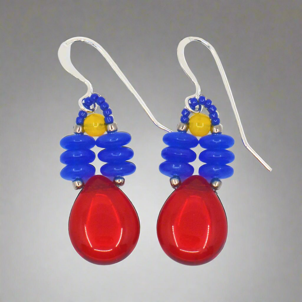 A pair of brightly colored earrings against a gray background. The earrings have a big clear red teardrop suspended from two parallel stacks of blue discs.