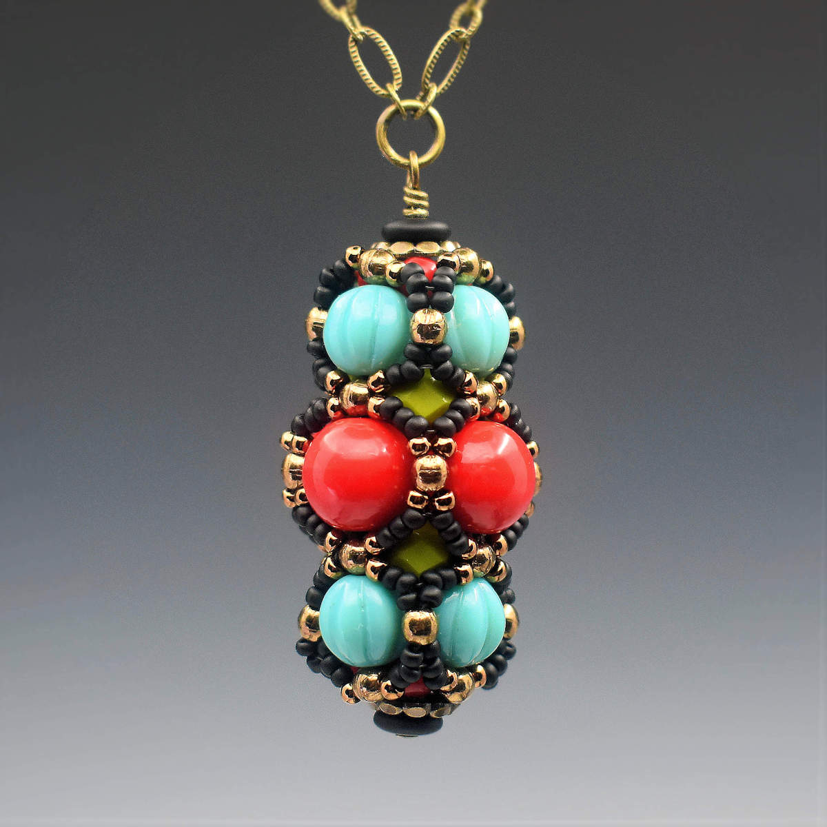 A turquoise and red pendant on brass chain hangs in front of a gray backrgound. The pendant has a central row of large red beads with a row of turquoise beads on either side. A netting of black and gold seed beads outlines all of the red and turquiose beads.