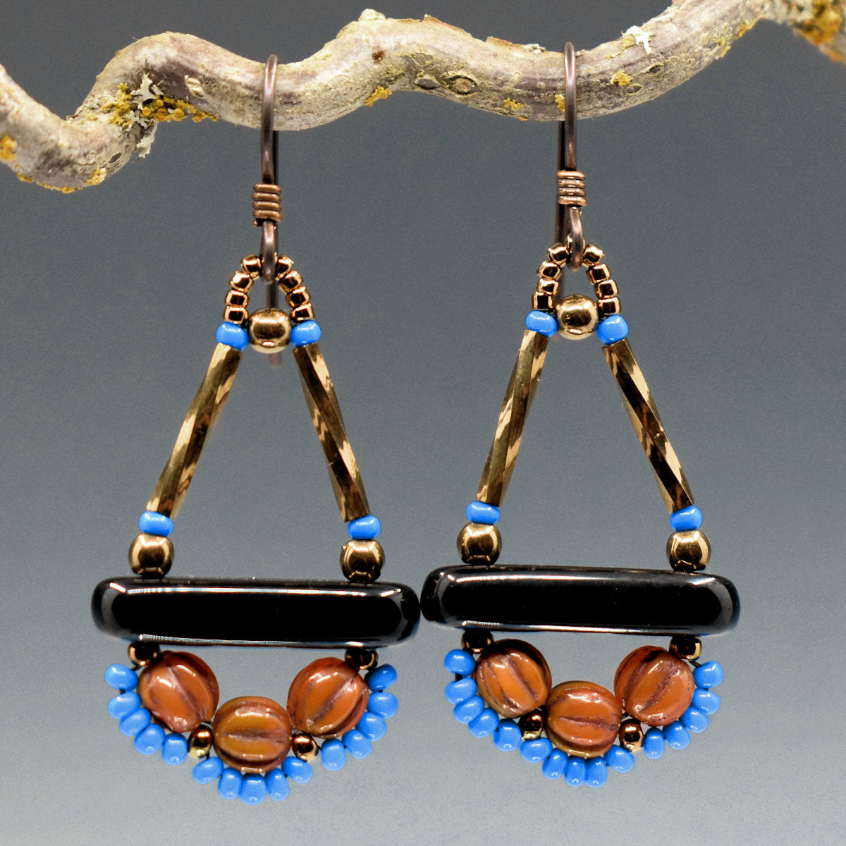 Gold, black, blue and brown earrings hang from a twisted twig. The earrings are a triangle on top, with gold tubes for upright sides and a black bar at the bottom. Below the bar there are three rust colored ridged round beads, each with a row of tiny blue seed beads below it.