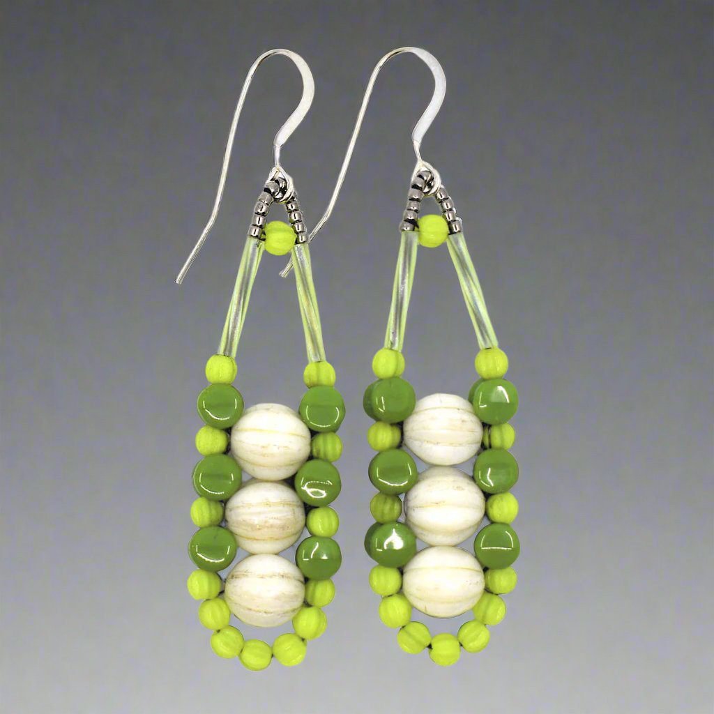 Cream and green earrings with silver accents and ear wires on a gray background. These earrings have three cream rounds stacked vertically like peas and held in place by an outline of bright chartreuse and darker avocado green beads.