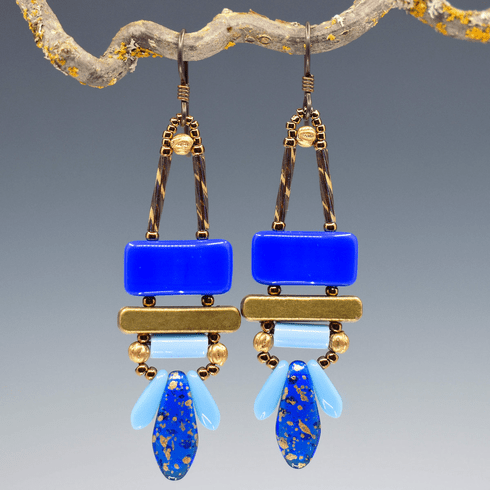 Blue and gold earrings with dark ear wires hang from a lichen covered twig against a gray background. These earrings have blue rectangles above gold bars. Below the bars are light blue tubes, and below a wide gold-speckled transparent blue dagger is sandwiched between two smaller light blue daggers.