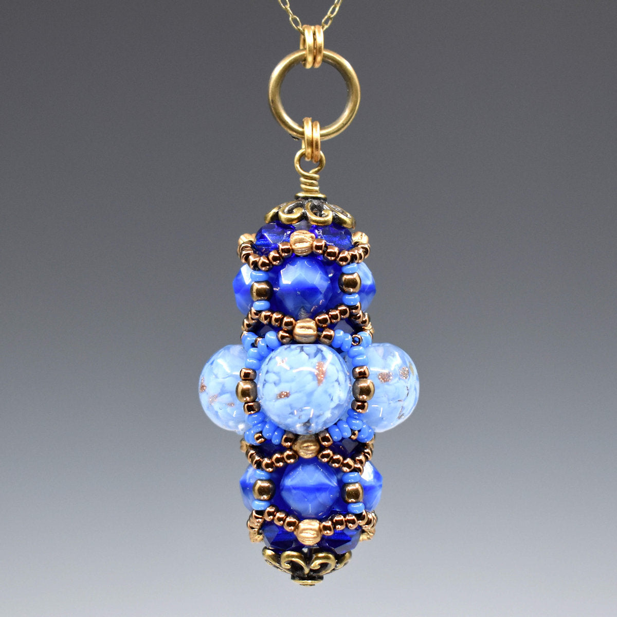 A blue and gold pendant hanging from a gold colored chain against a gray background. The middle of the pendant has clear beads filled with flecks of light blue and some gold glitter accents. The top and bottom layers are made from smaller medium blue beads. The whole pendant is overlaid with gold and blue seed beads.