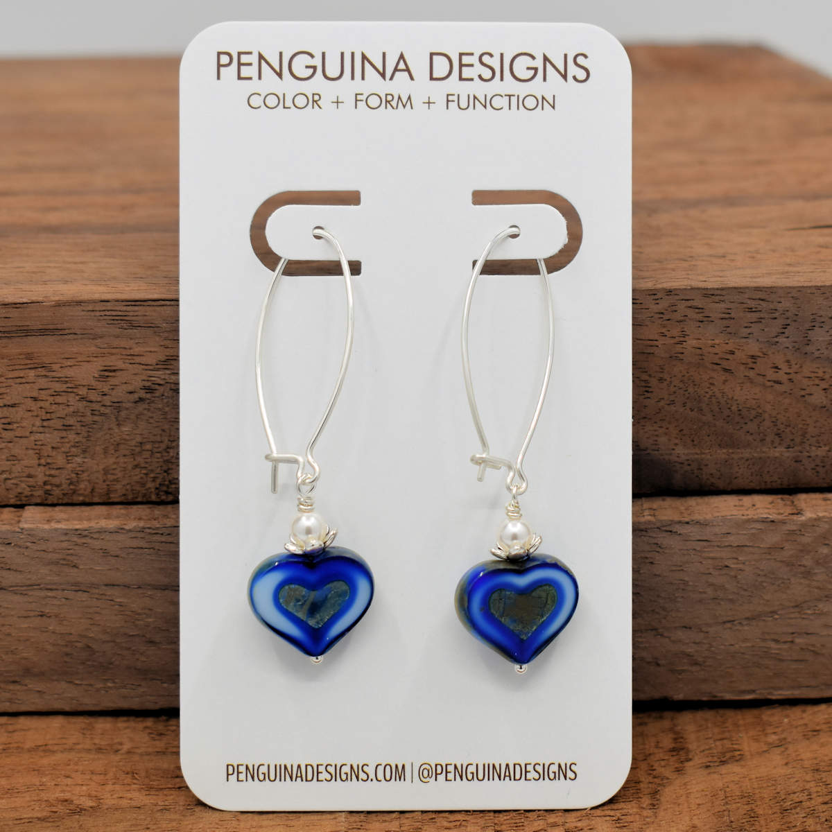 A pair of earrings on a white card rest against a wood background. The earrings have long silver oval wires that latch and blue hearts at the bottom with a white pearl bead. The blue hearts are a variegated dark and light blue and have a darker stonelike texture heart pressed into the center.