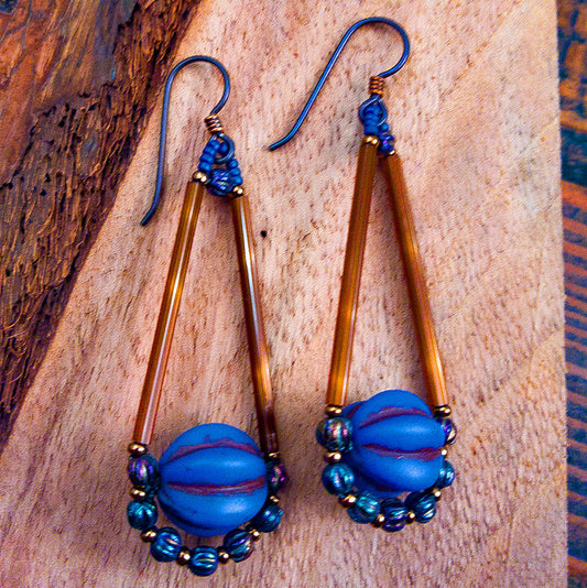 A pair of brown and cobalt blue teardrop shaped earrings lay on wood. The teardrop shape of the earrings is formed with two long brown beads on the upper, straight sides, and at the bottom is a matte blue bead with deep horizontal ridges. The bottom of the teardrop is outlined by small iridescent blue rounds alternating with gold seed beads. The ear wires are dark metal.