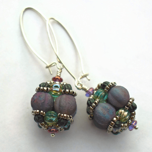 A pair of earrings laying against a white background. The earrings have long silver oval wires that latch and there is a beaded ball dangling from the bottom of each earring. The beaded beads are s made from rough textured lilac beads surrounded by layers of silver and sea green beads.
