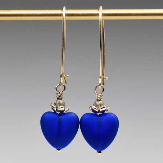 A pair of earrings hand from a gold bar, against a gray background. The earrings have long silver oval wires that latch and have drops that are made from small frosted cobalt glass hearts topped by a flower shaped cup that holds a silvery bead.