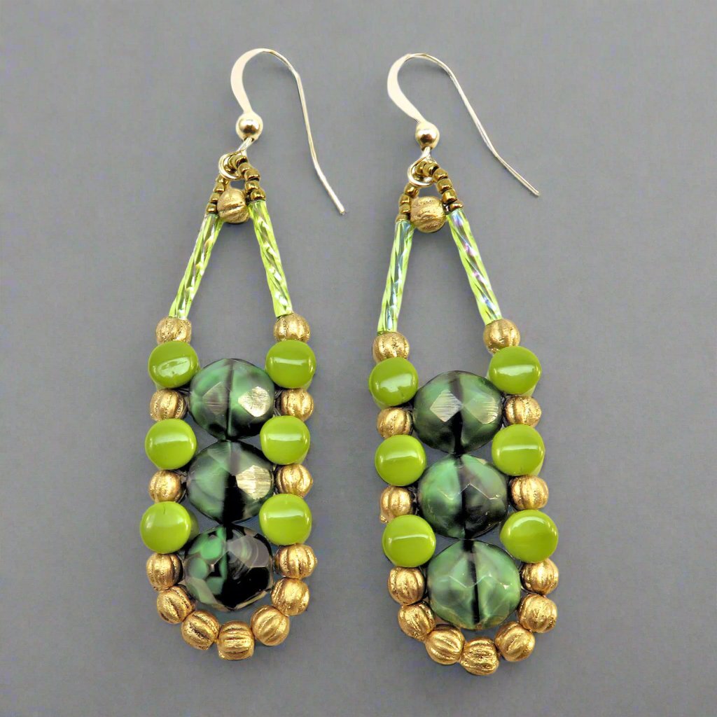 Long earrings in different shades of green with gold accents and ear wires on a gray background. There are three swirled medium green beads stacked vertically in the center of these beads, like peas, surrounded by an outline of gold melon and avocado pellet beads.