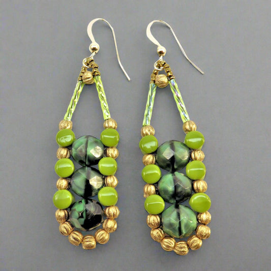 Long earrings in different shades of green with gold accents and ear wires on a gray background. There are three swirled medium green beads stacked vertically in the center of these beads, like peas, surrounded by an outline of gold melon and avocado pellet beads.