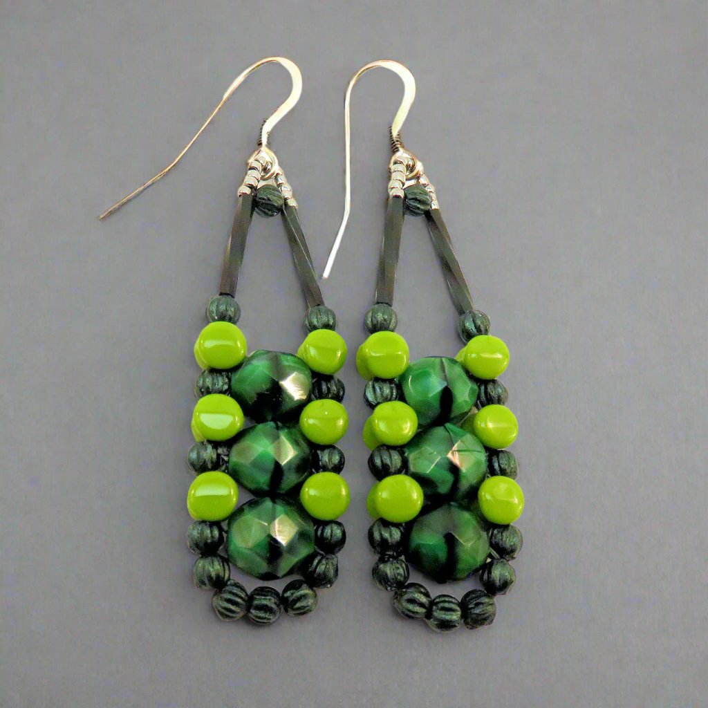 Long earrings in different shades of green with silver accents and ear wires on a gray background. There are three swirled medium green beads stacked vertically in the center of these beads, like peas, surrounded by an outline of dark metallic green and avocado green beads.