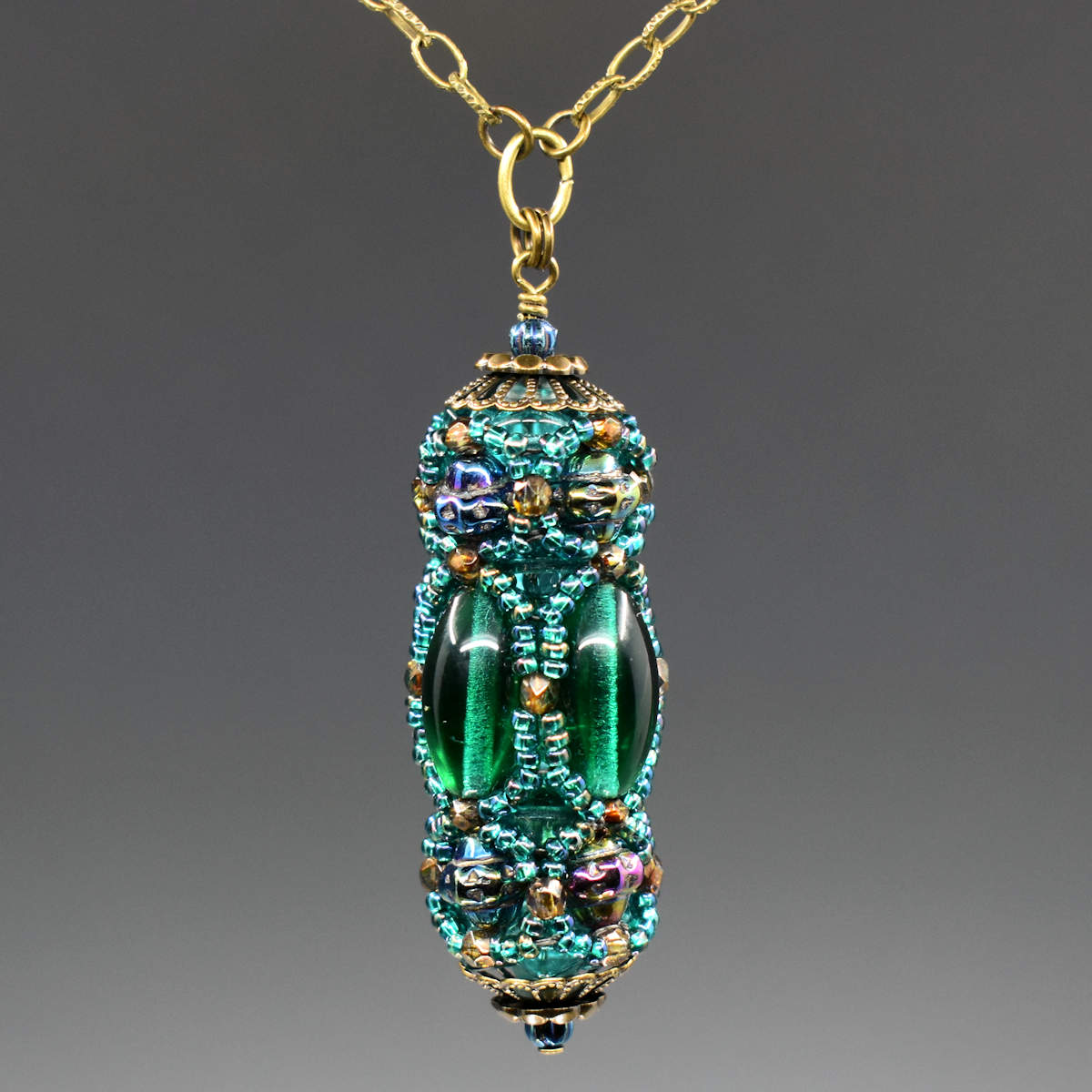 A turquoise and green beaded pendant hangs from a gold colored chain. The pendant has long oval beads that form the center row and textured iridescent beads that form a decorative top and bottom row. A netting of translucent peacock green seed beads is overlaid, outlining all of the larger beads.